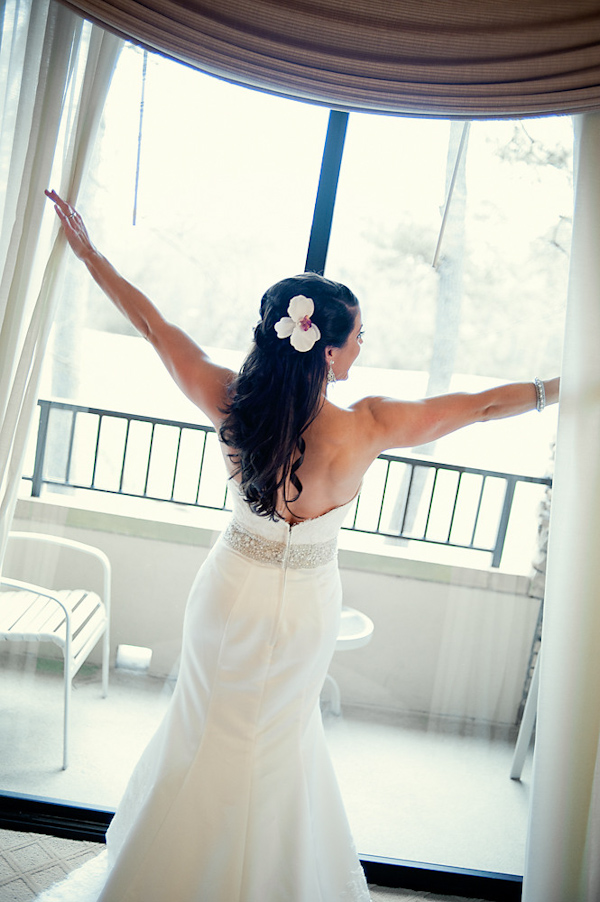 Beautiful bride wearing white mermaid style dress with a white flower in her hair pushing curtains back to look out the window - photo by Houston based wedding photographer Adam Nyholt